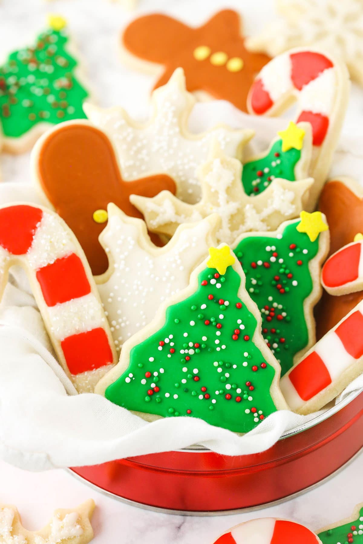 Sugar cookies shaped like snowflakes, candy canes and Christmas trees inside of a metal tin on a kitchen countertop.