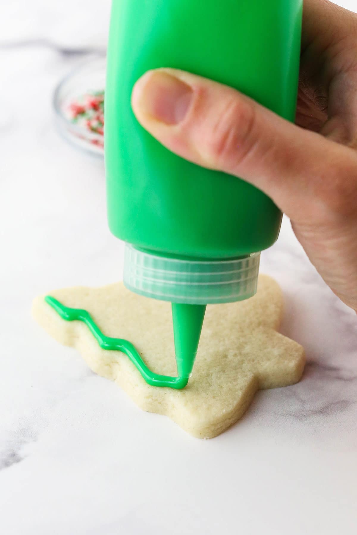 Green icing being added to a small sugar cookie shaped like a Christmas tree