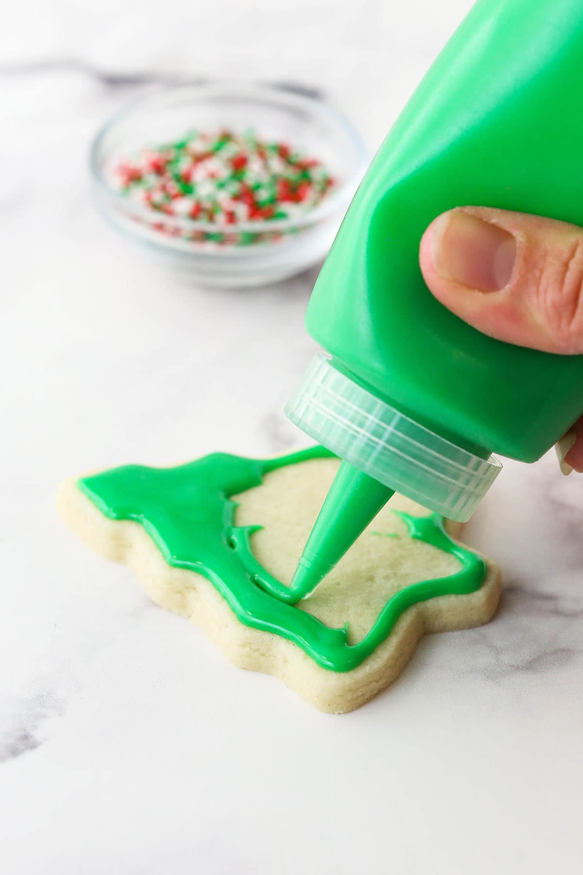 A hand squeezing a bottle of green icing onto a tree-shaped sugar cookie