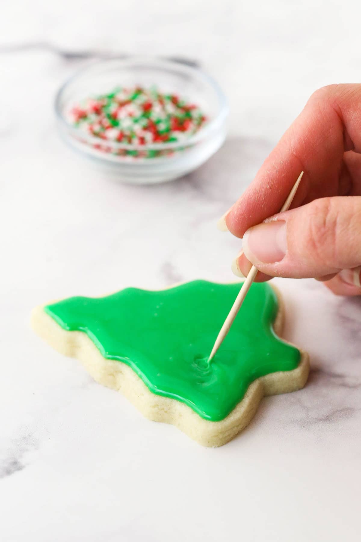 This glossy sugar cookie icing is easy to make in just 15 minutes! It hardens beautifully on top of cookies and tastes as good as it looks. A toothpick smoothing out a layer of green icing on top of a sugar cookie