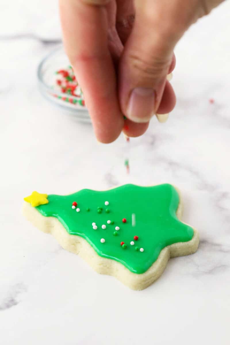 This glossy sugar cookie icing is easy to make in just 15 minutes! It hardens beautifully on top of cookies and tastes as good as it looks. Little round sprinkles being dropped onto a sugar cookie decorated like a Christmas tree