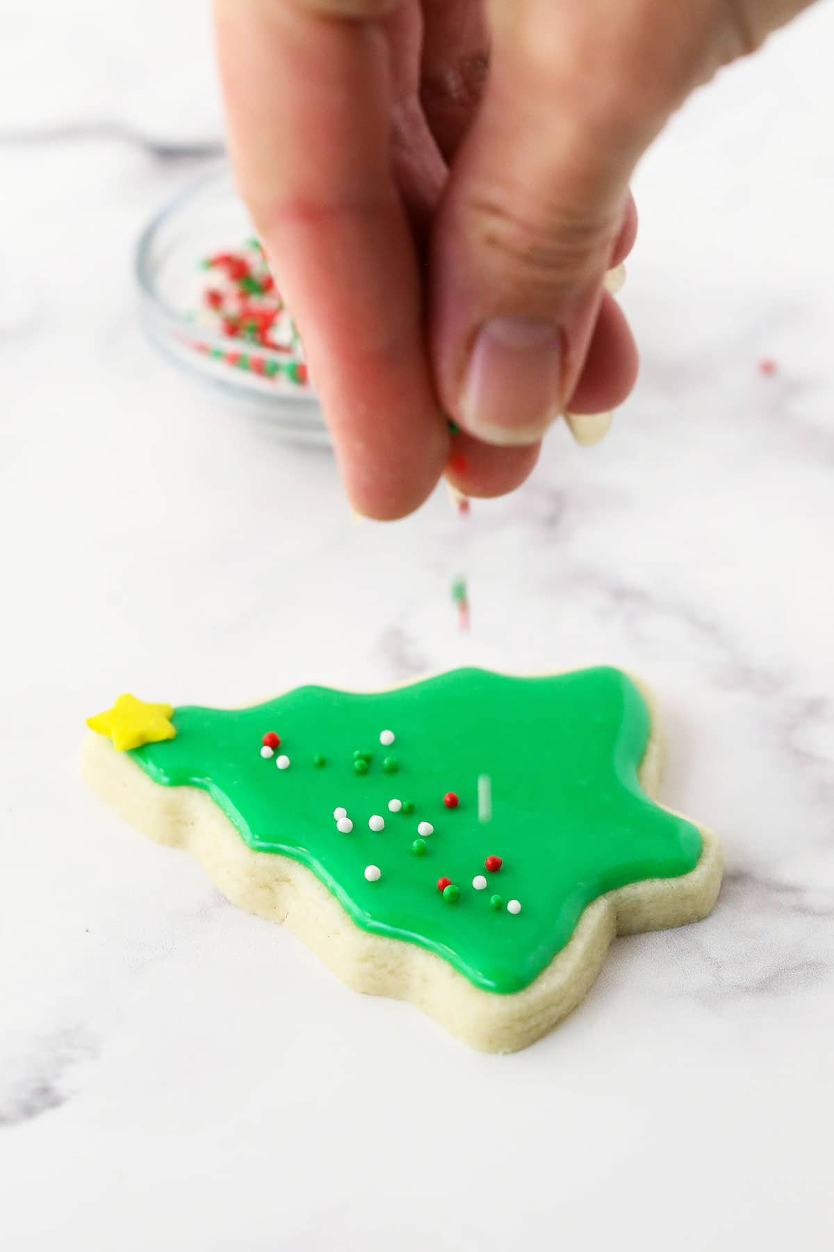 Little round sprinkles being dropped onto a sugar cookie decorated like a Christmas tree