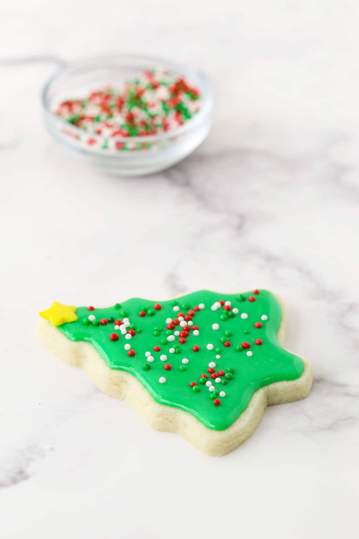 A cut-out Christmas tree sugar cookie with icing and sprinkles on a marble countertop with a bowl of sprinkles in the background