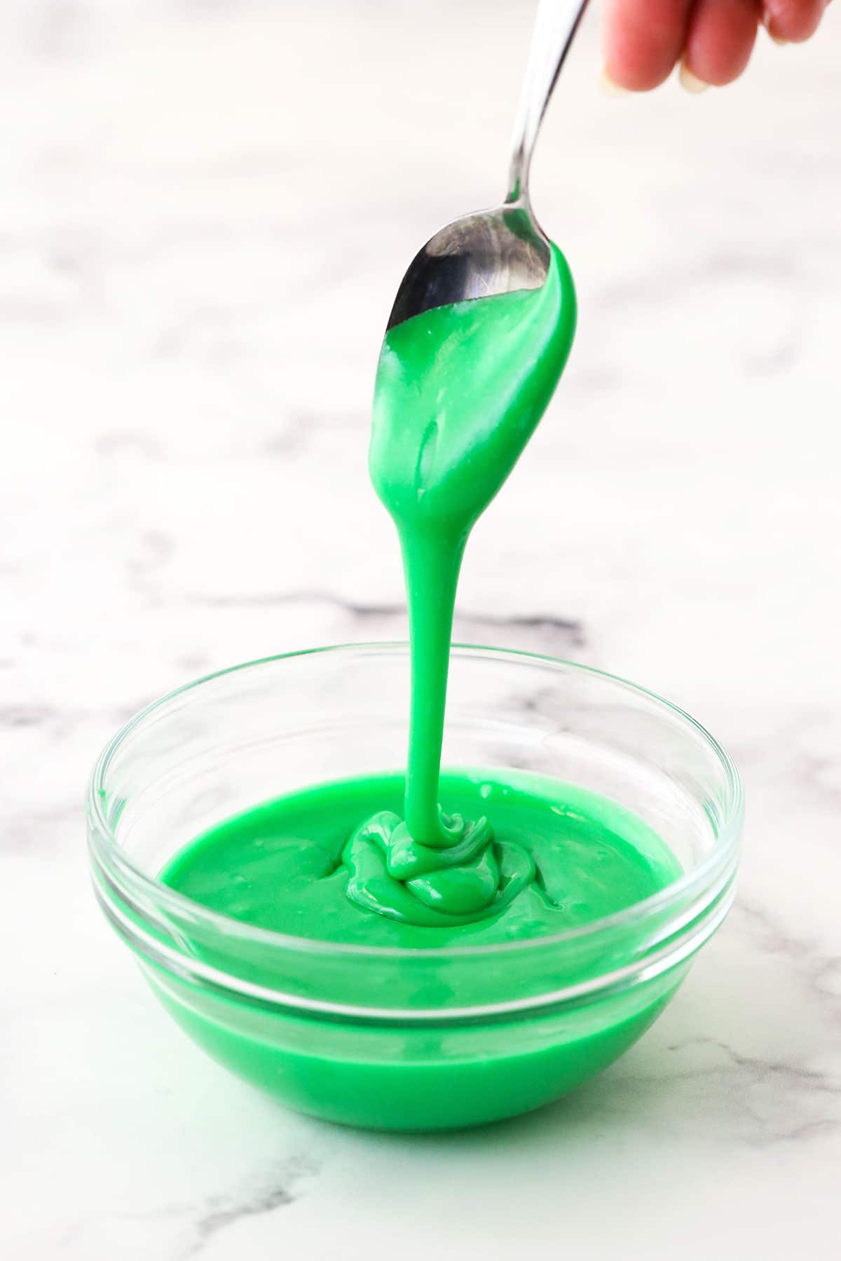 A spoon drizzling some green icing into a small glass bowl with more icing in it