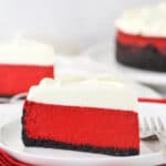 A piece of red velvet cheesecake on a plate with another piece and the remaining cheesecake behind it