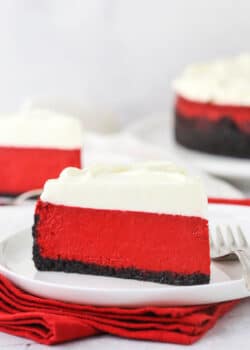 A piece of red velvet cheesecake on a plate with another piece and the remaining cheesecake behind it