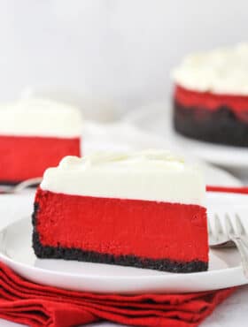 A piece of red velvet cheesecake on a plate with another piece and the remaining cheesecake behind it