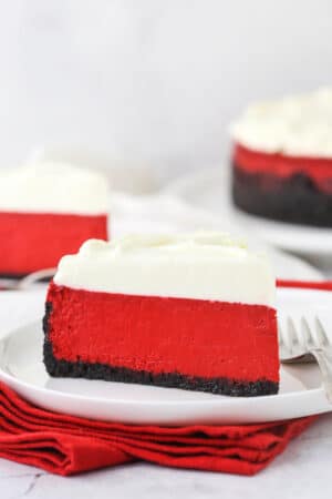 A piece of red velvet cheesecake on a plate with another piece and the remaining cheesecake behind it