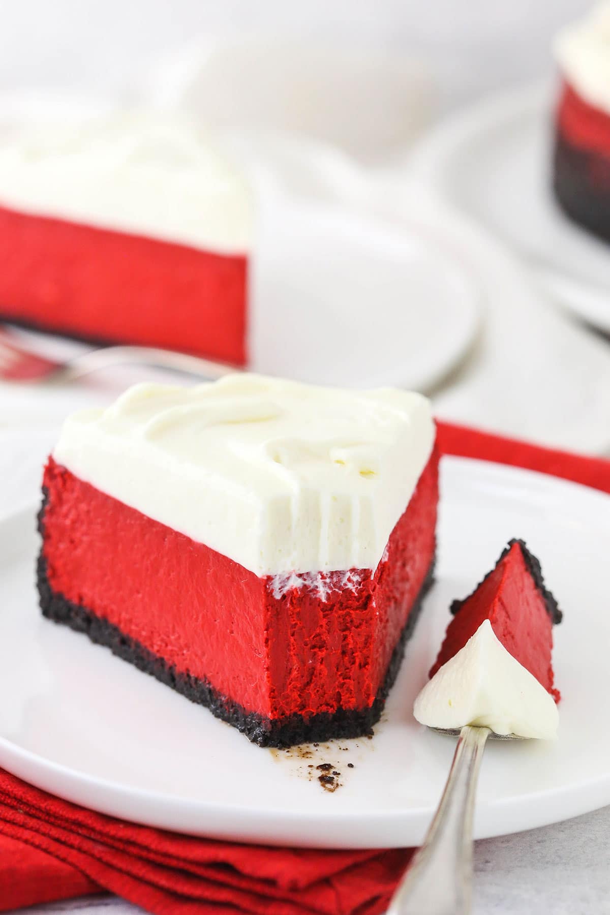 A slice of red velvet cheesecake on a small plate with one bite on a fork