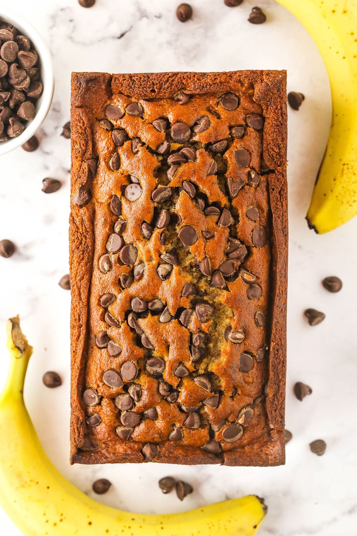 A loaf of chocolate chip banana bread on a countertop with two bananas and a bowl of chocolate chips