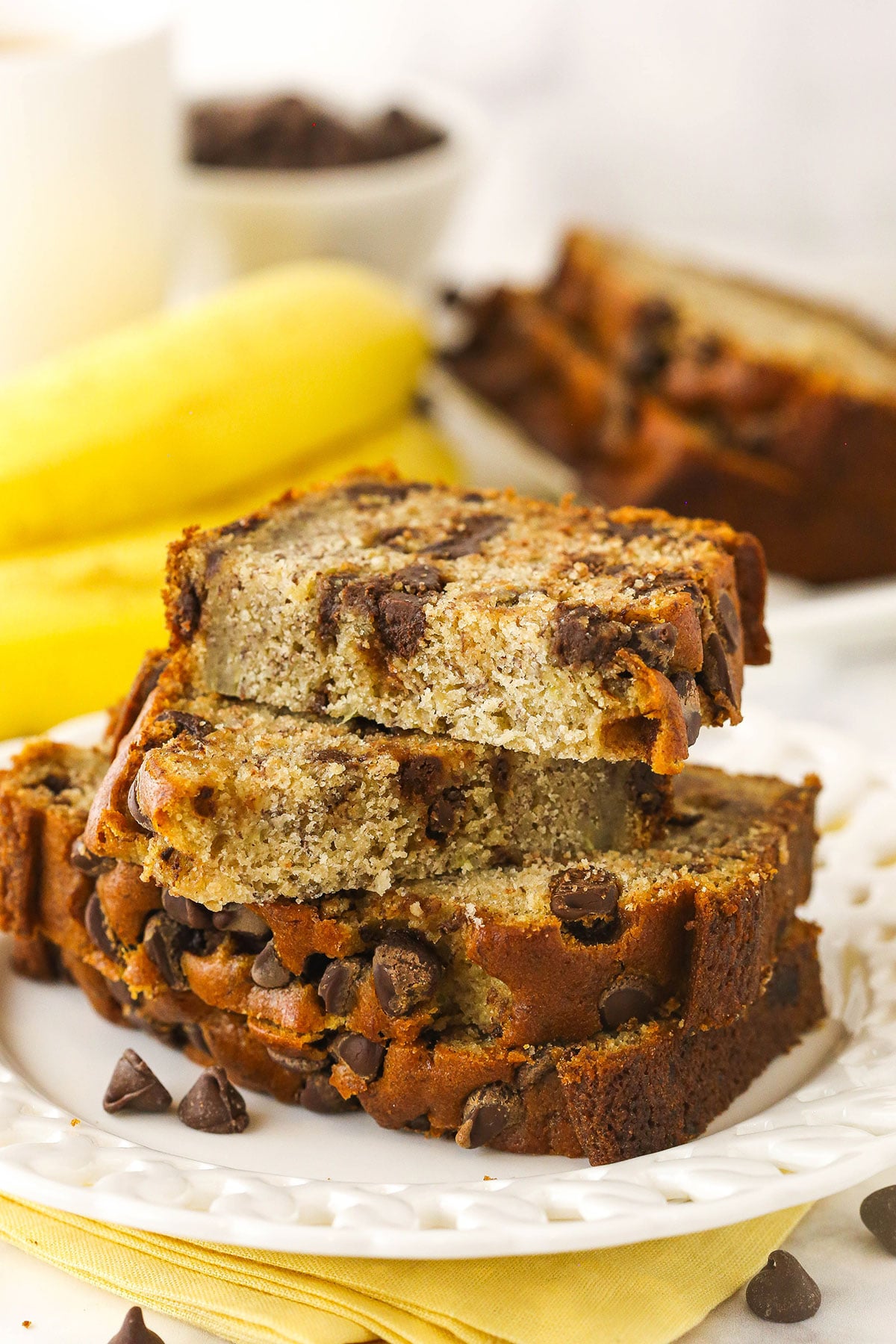 Slices of chocolate chip banana bread stacked onto a plate with two bananas in the background