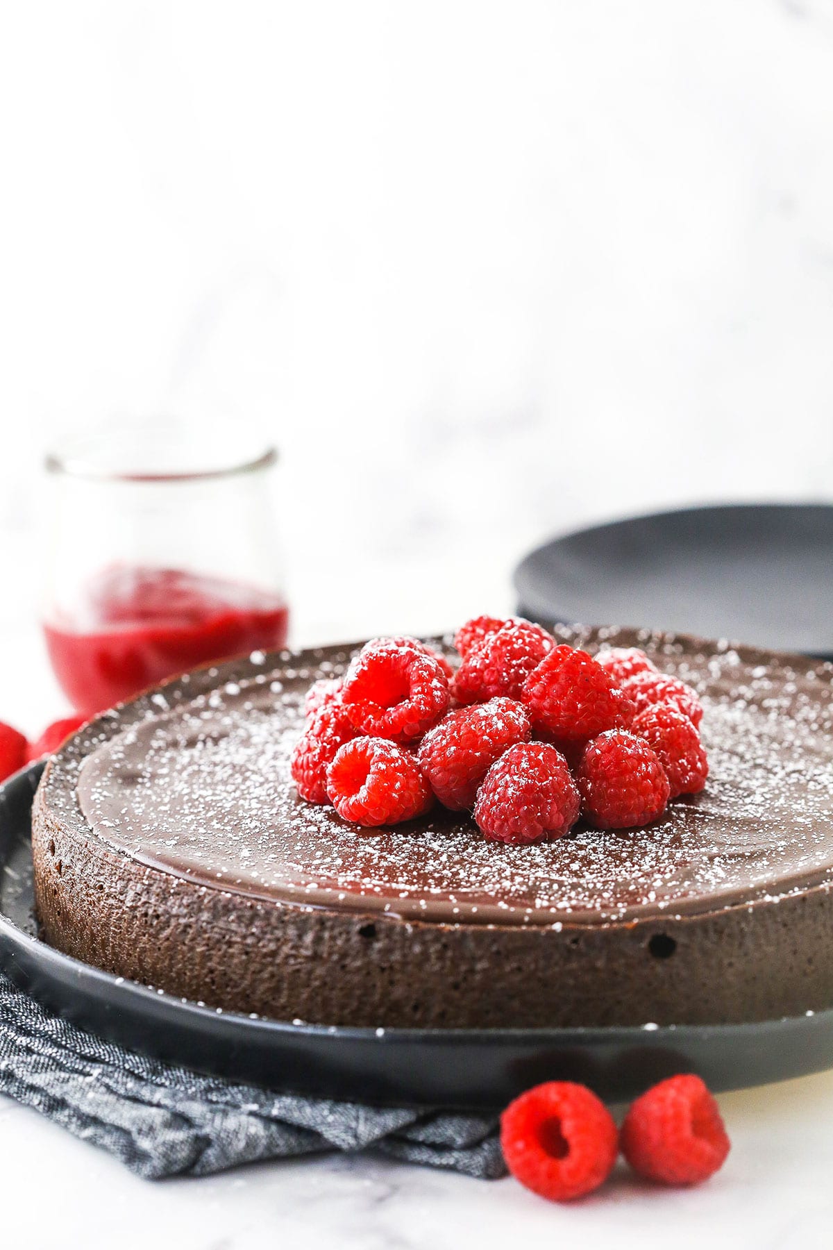 A flourless chocolate cake on a platter with two raspberries in the foreground and a plate in the background