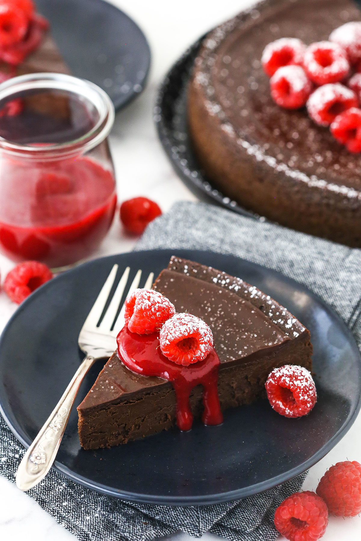 A slice of chocolate torte on a plate with raspberry sauce and the remaining cake in the background