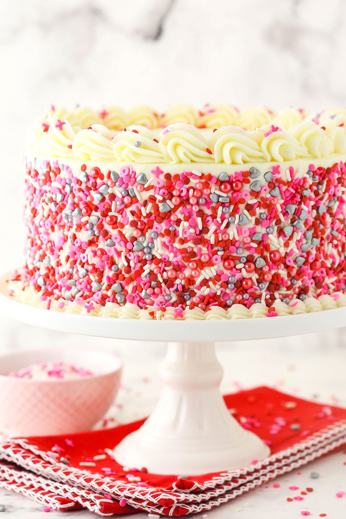 A cake stand holding a completed red velvet layer cake with a red kitchen towel underneath it