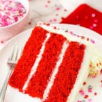 A slice of homemade red velvet cake on a plate with a bowl of sprinkles behind it