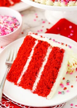 A slice of homemade red velvet cake on a plate with a bowl of sprinkles behind it