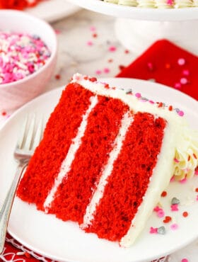 A slice of homemade red velvet cake on a plate with a bowl of sprinkles behind it