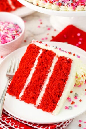 A slice of homemade red velvet cake on a plate with a bowl of sprinkles behind it