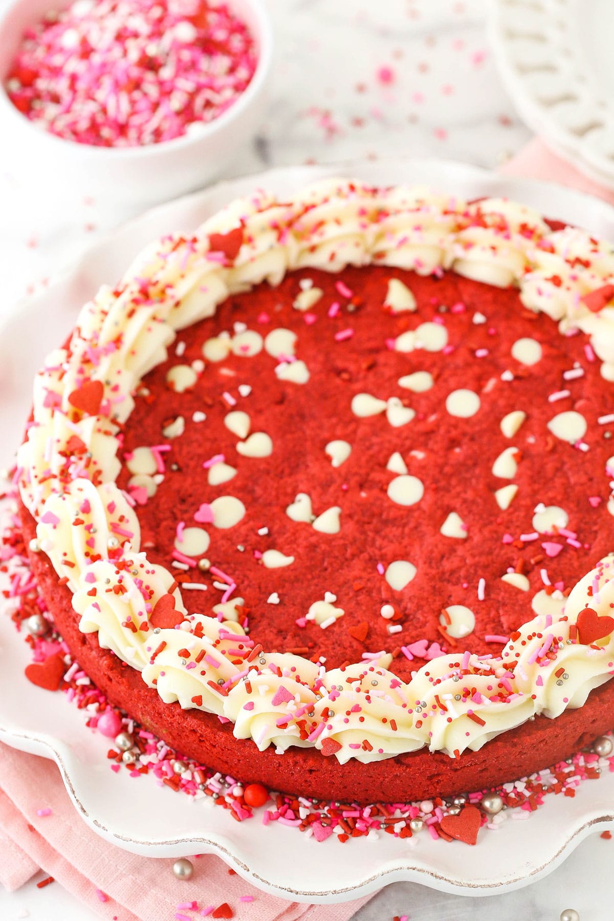 A red velvet cookie cake with white chocolate chips and a rim of cream cheese frosting with a bowl of sprinkles behind it