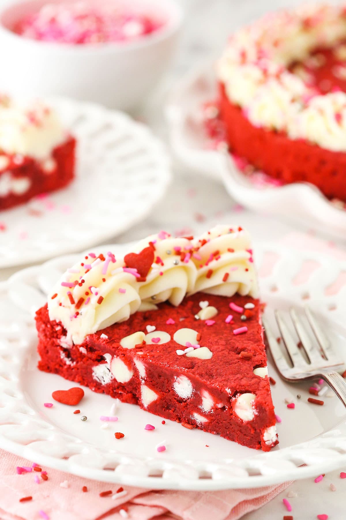 A frosted slice of chocolate chip cookie cake on a plate with red and pink sprinkles on top of the cream cheese frosting