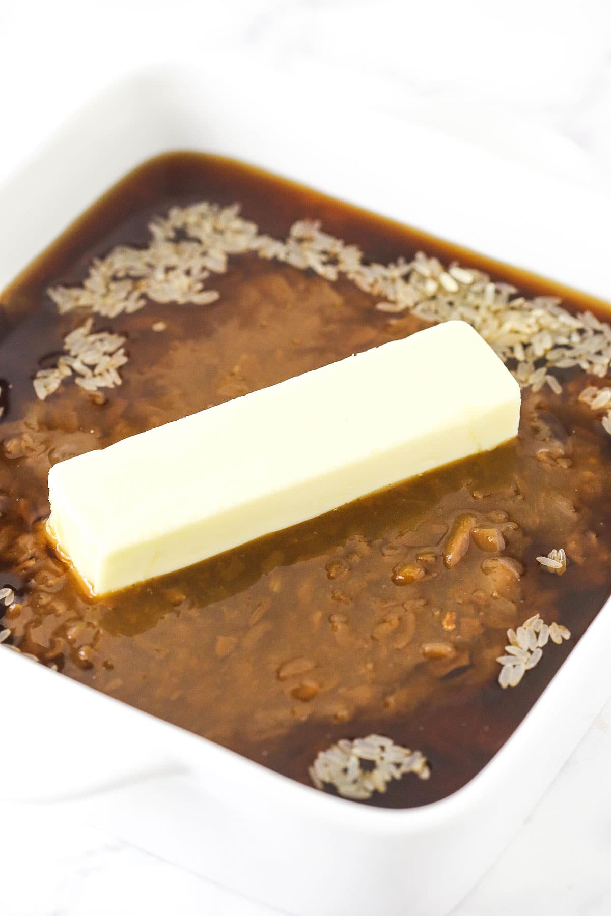 A baking dish full of uncooked brown rice, condensed soups and a stick of butter