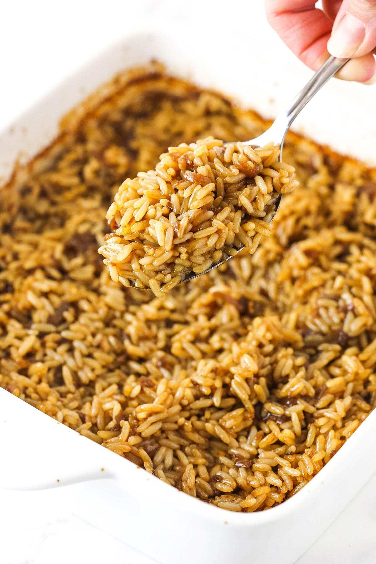 A metal spoon holding up a big bite of baked rice with a pan full of more rice beneath it