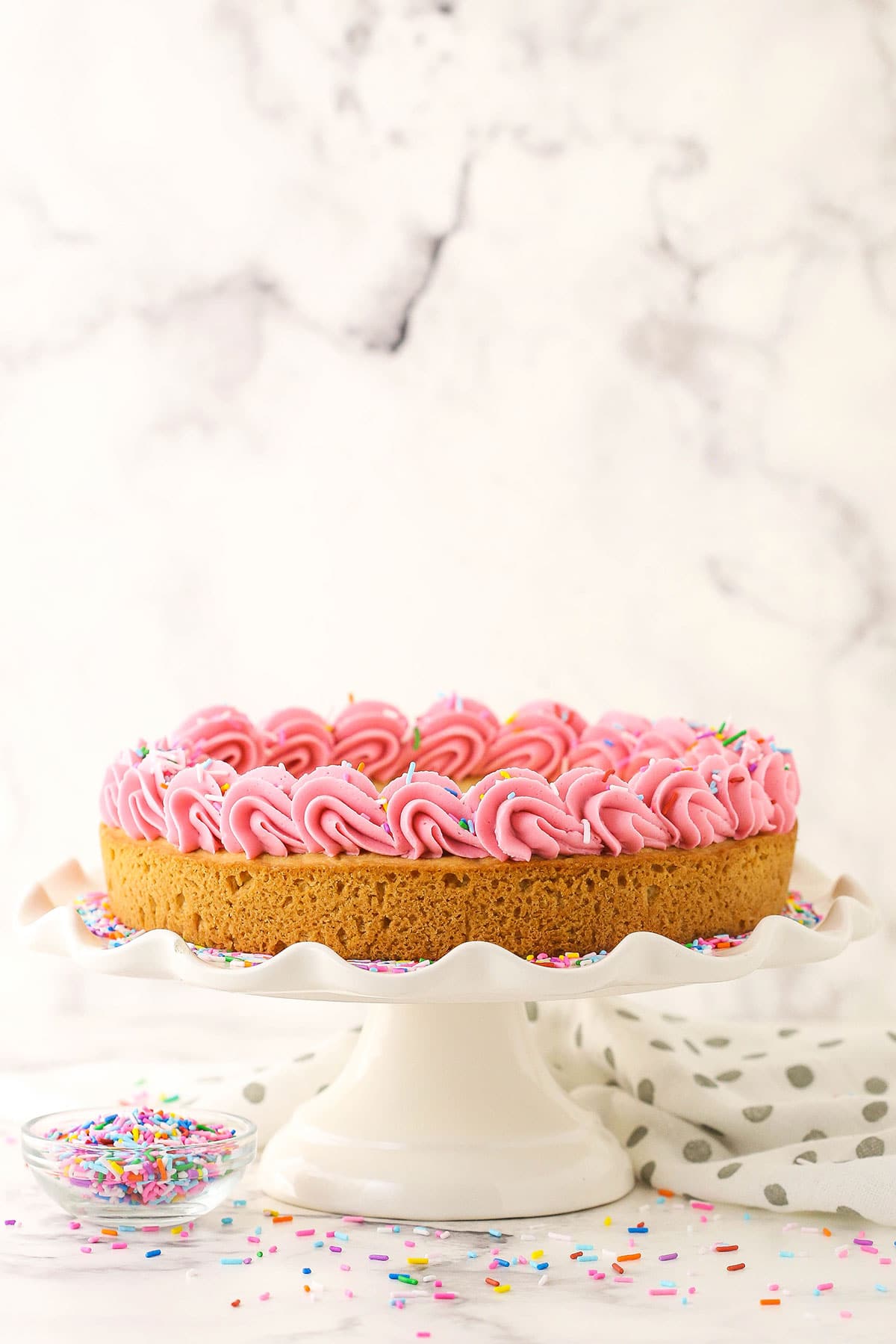 Side view of a cookie cake topped with piped pink frosting, on a white cake plate.