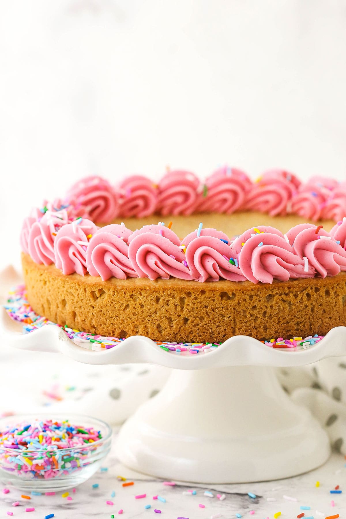 A vanilla cake on a white cake plate. The cake is decorated with pink frosting and multicolored sprinkles.