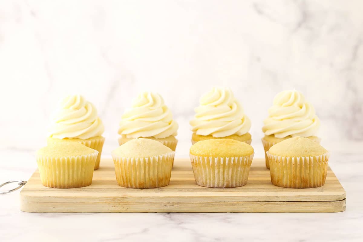 Four unfrosted cupcakes on a cutting board with a frosted cupcake directly behind each one