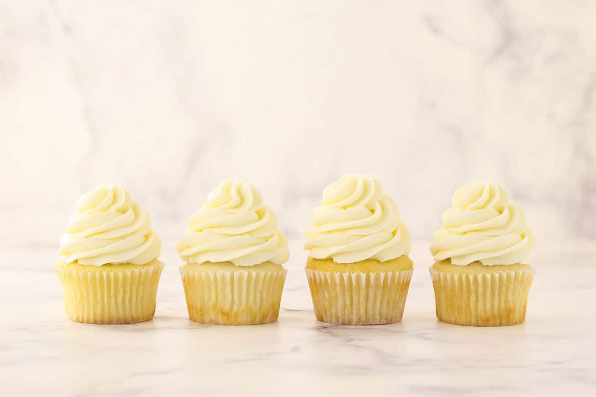 Four frosted vanilla cupcakes on a marble countertop with a matching marble backdrop
