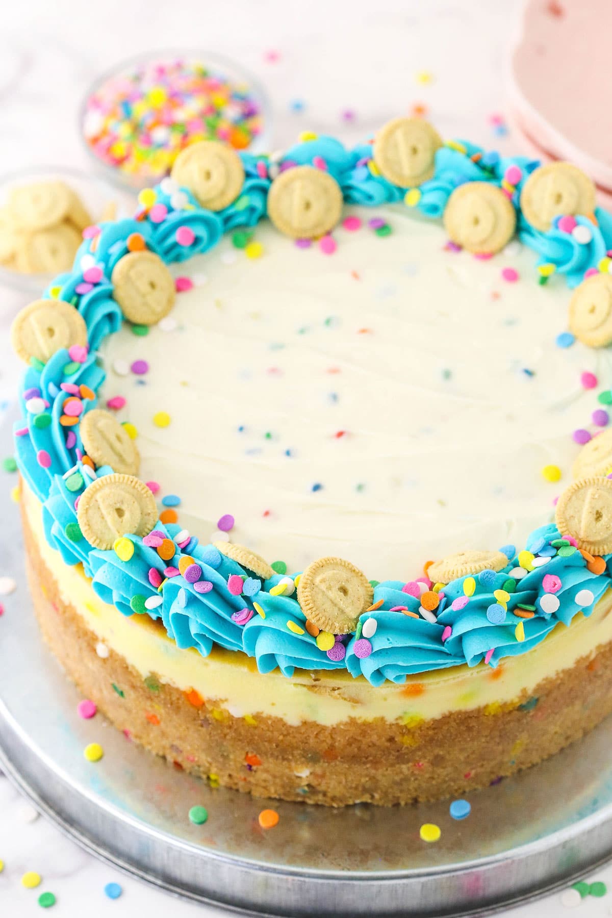 A Dunkaroos cheesecake on a large metal serving platter with bowls of sprinkles and Dunkaroo cookies behind it