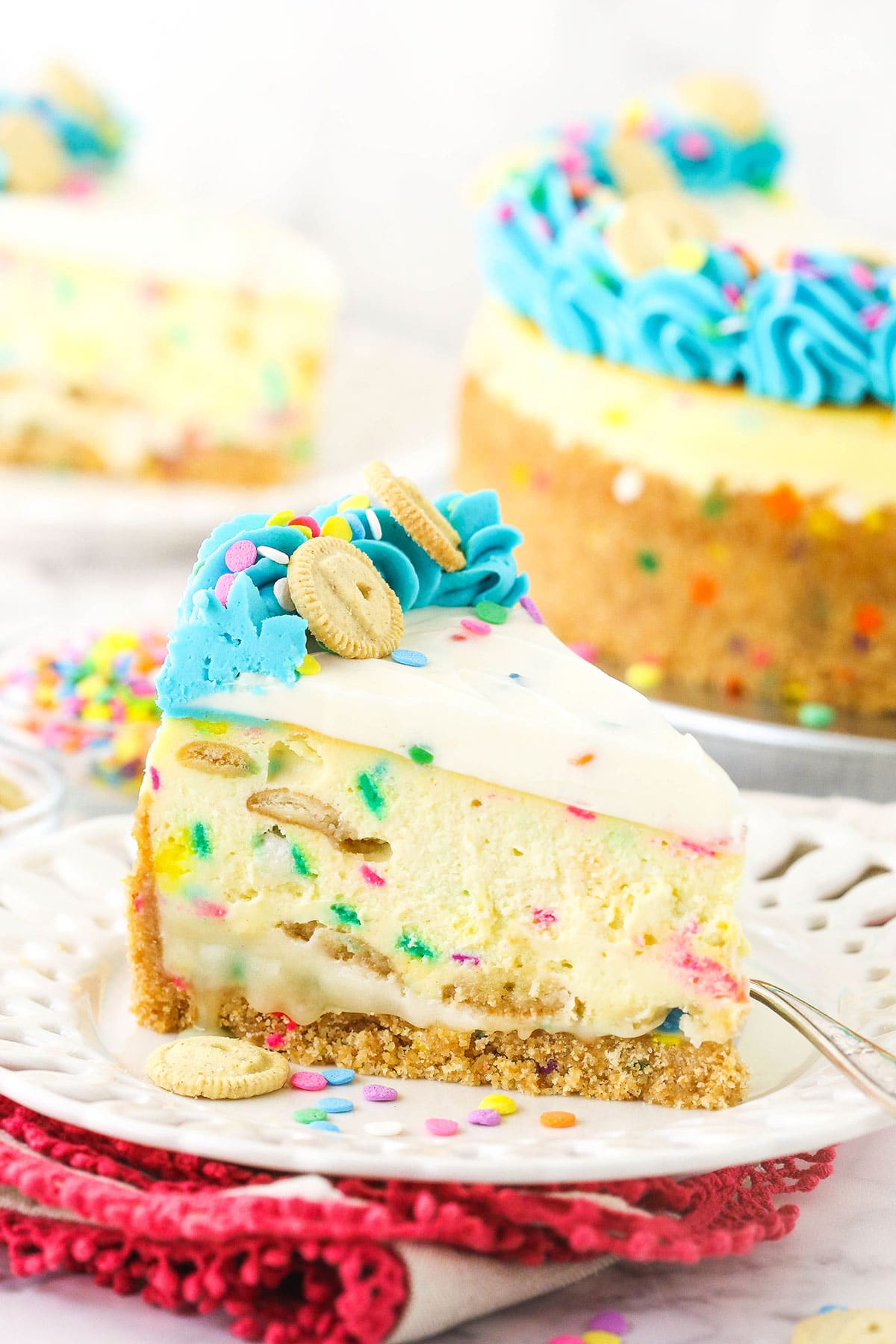 A slice of confetti frosting cheesecake on a plate with a bowl of sprinkles behind it