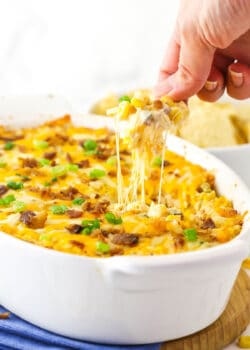 A chip that has just been dunked into a dish of corn dip and is being pulled out with gooey cheese sticking from the chip to the top of the dip