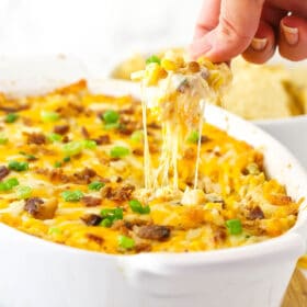 A chip that has just been dunked into a dish of corn dip and is being pulled out with gooey cheese sticking from the chip to the top of the dip