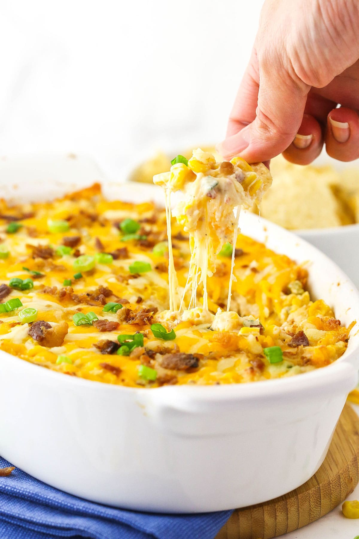 A chip that has just been dunked into a dish of corn dip and is being pulled out with gooey cheese sticking from the chip to the top of the dip