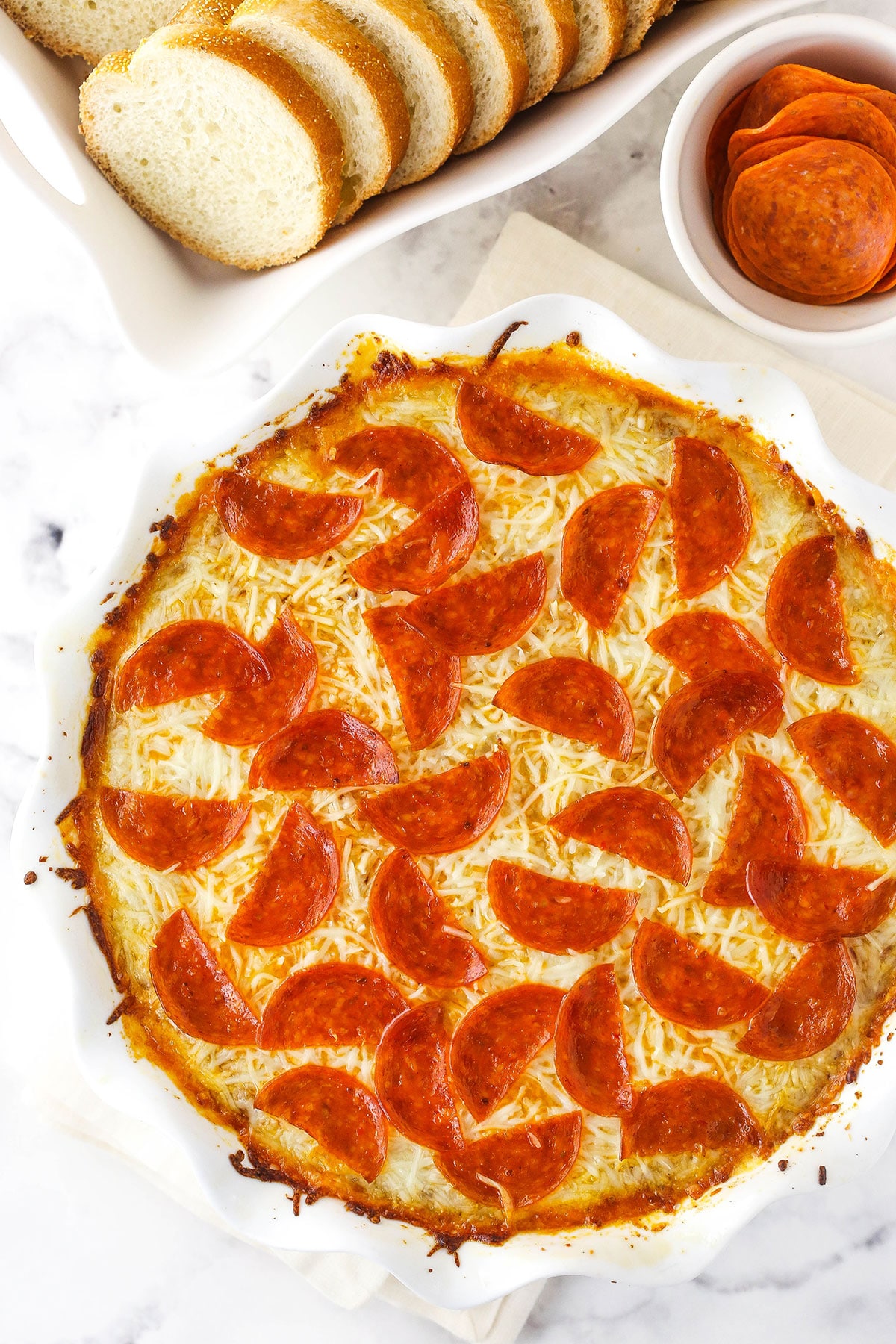 Overhead shot of a round baking dish of pizza dip, with marinara and sliced baguettes nearby.