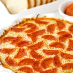 Pizza dip in a white dish, with a white dish of sliced French bread and a cup of marinara.