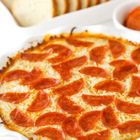 Pizza dip in a white dish, with a white dish of sliced French bread and a cup of marinara.