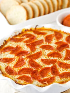 Pizza dip in a white dish, with a white dish of sliced French bread and a cup of marinara.