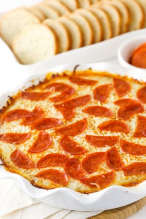 Pizza dip in a white dish, with a white dish of sliced French bread and a cup of marinara.