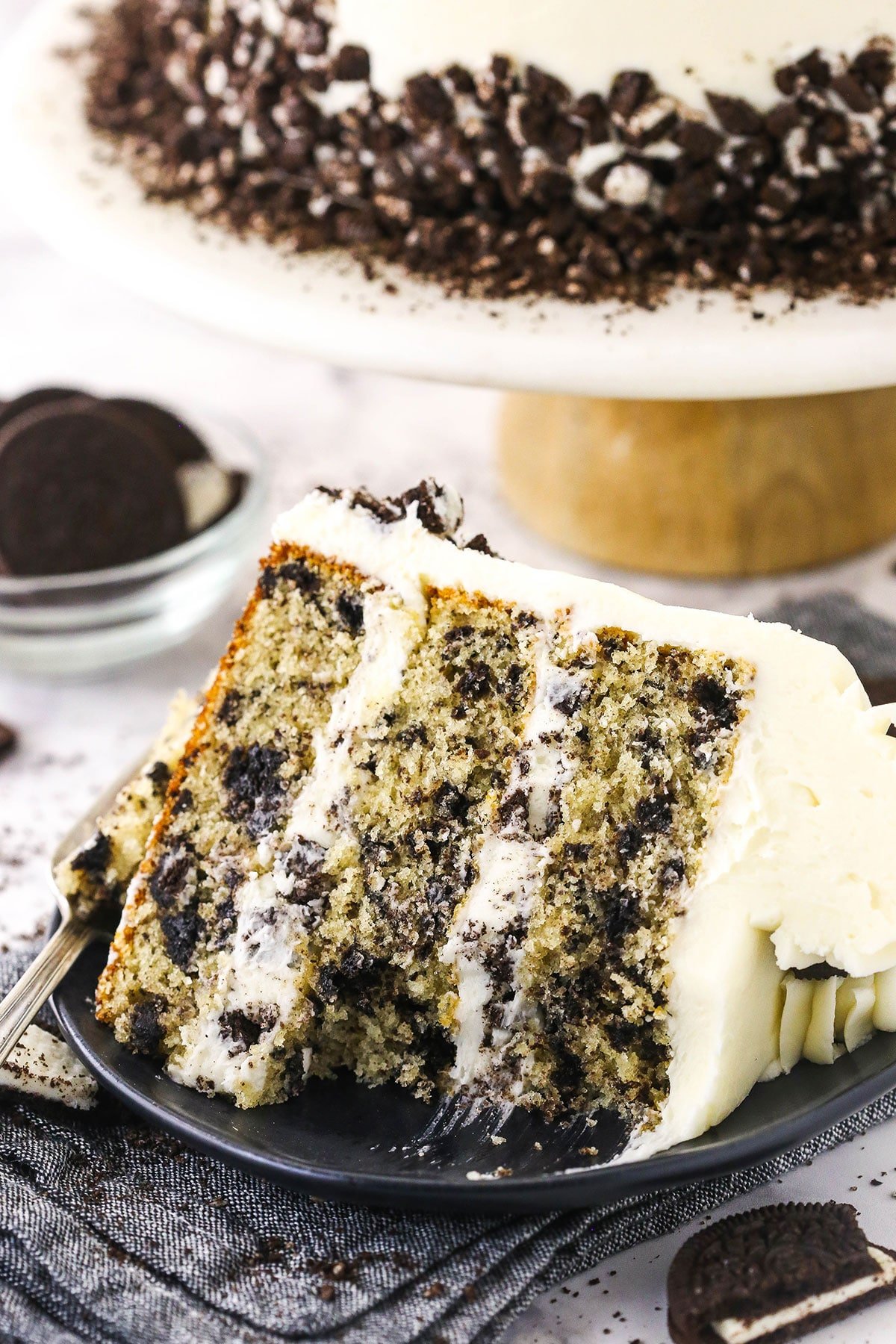 A half-eaten slice of Oreo cake on a plate with the rest of the cake on a stand behind it