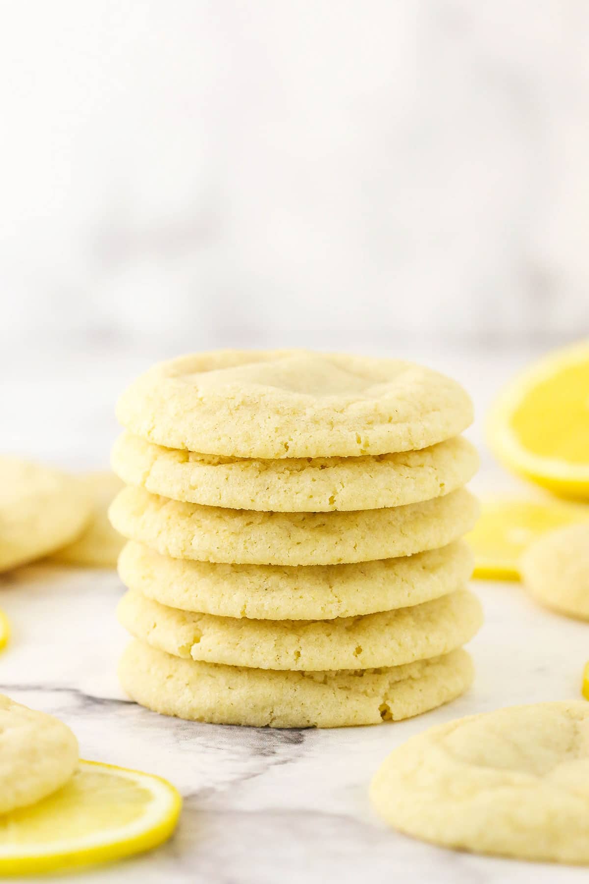 Six lemon sugar cookies stacked on a marble countertop with more cookies surrounding them