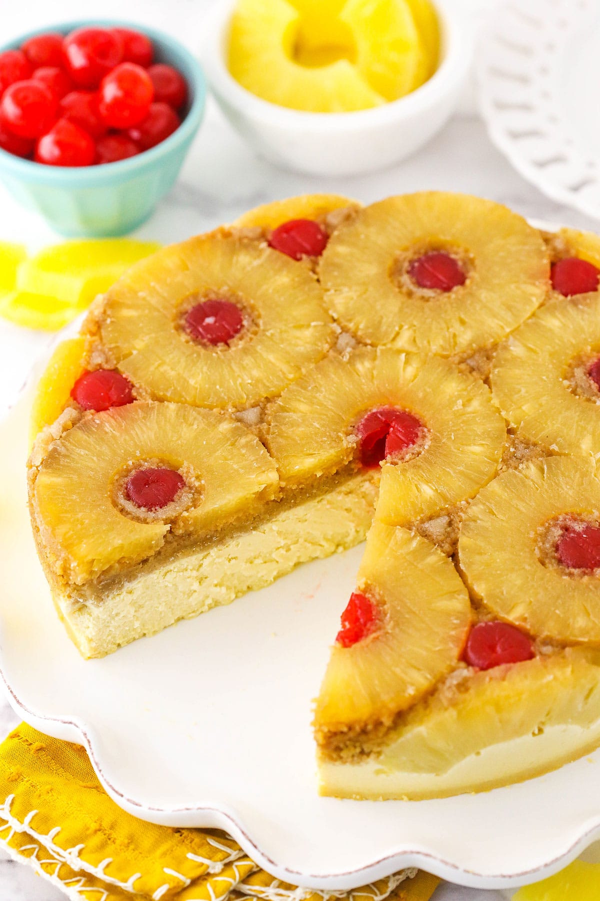 A cheesecake with one slice cut out. Pineapple and cherries are on the table near the cake.