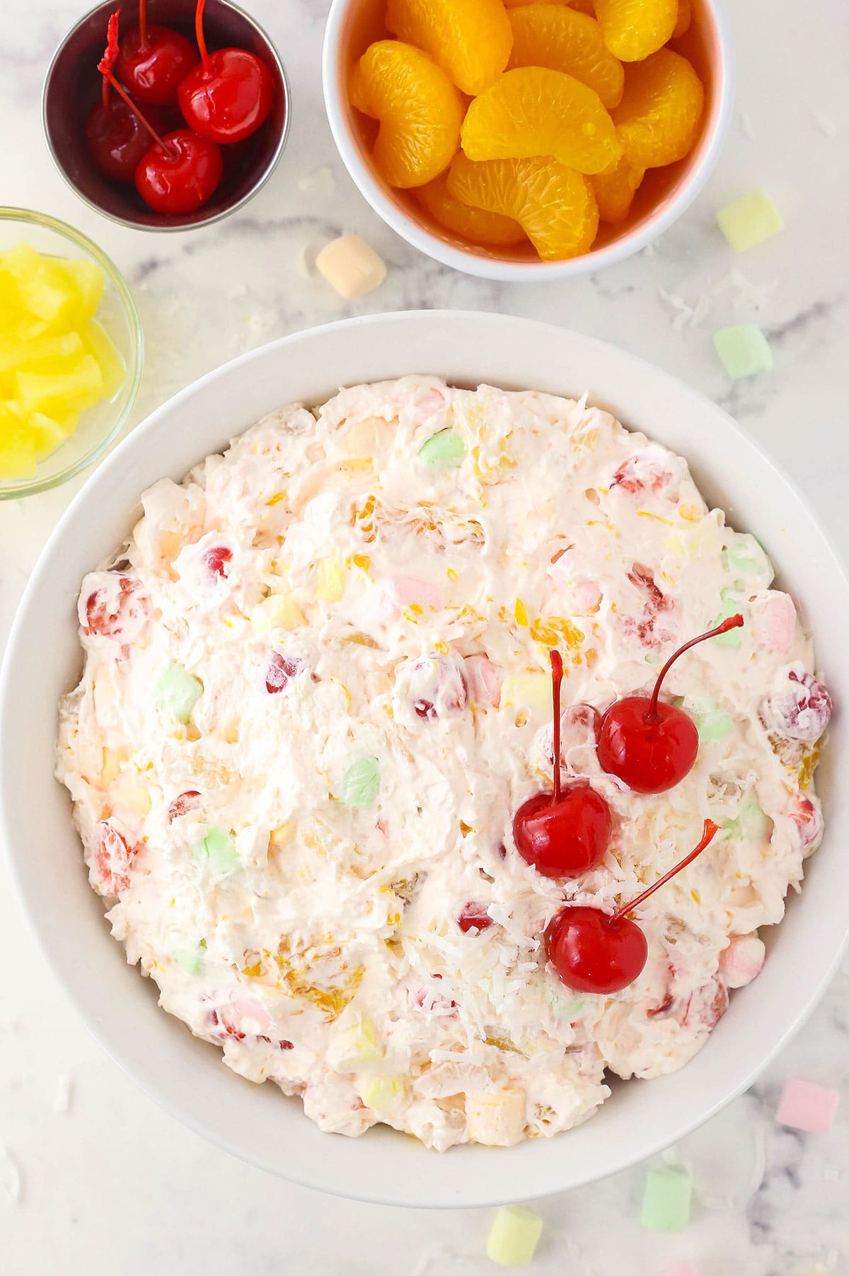 An ambrosia salad in a big bowl on top of a granite surface with fruits and mini marshmallows beside it