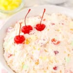 A large serving bowl full of marshmallow fruit salad with pineapple chunks, cherries and tangerines behind it