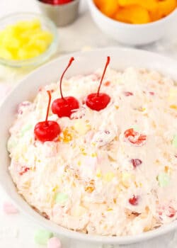 A large serving bowl full of marshmallow fruit salad with pineapple chunks, cherries and tangerines behind it