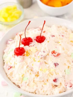 A large serving bowl full of marshmallow fruit salad with pineapple chunks, cherries and tangerines behind it