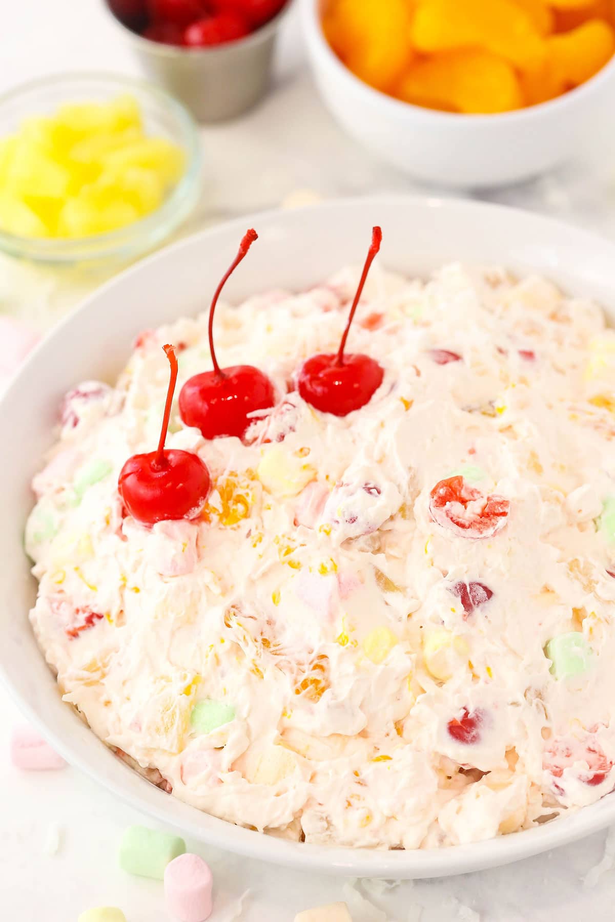 A large serving bowl full of marshmallow fruit salad with pineapple chunks, cherries and tangerines behind it