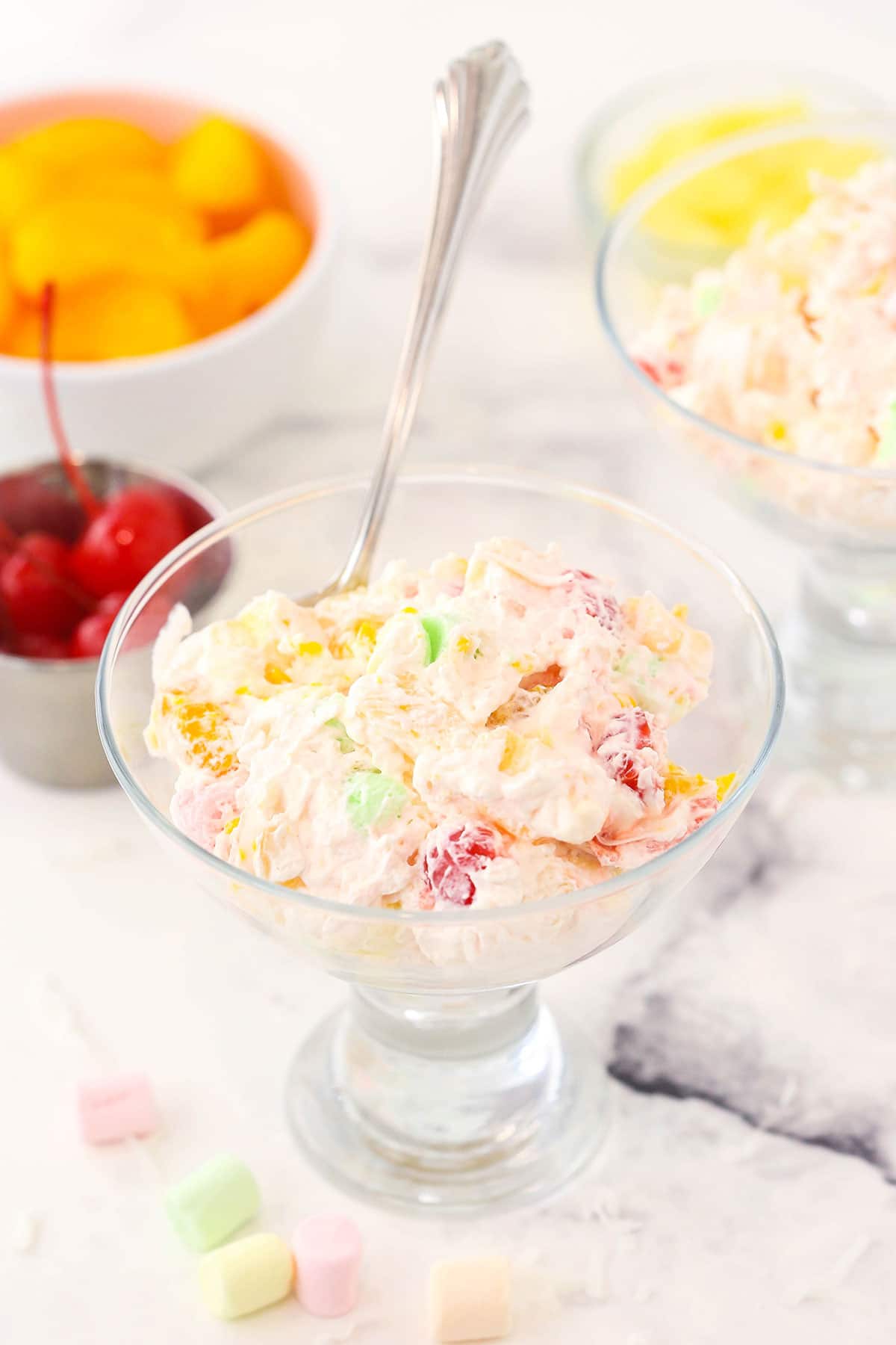 A serving of ambrosia salad inside of a cocktail glass with canned fruit in the background