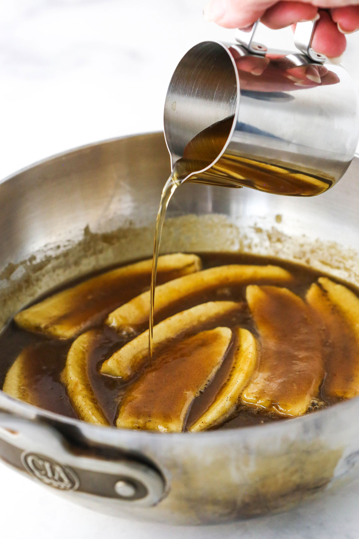 Three tablespoons of dark rum being poured over a skillet full of bananas foster